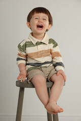 Wall Mural - Emotional little boy sitting on stool against light grey background