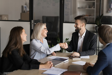 Canvas Print - Older and younger businesspeople, teammates shaking hands seated at desk in office take part in group briefing in modern meeting room. Shareholders greeting each other, start negotiations in boardroom