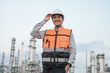 © PhugunStock - Asian male engineer smiling at camera, touching his helmet, wearing a safety uniform standing in front of an oil refinery with radio equipment. Ordering work at an oil refinery
