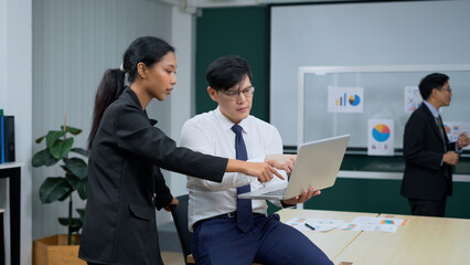 asian business man sitting on table holding laptop computer talking to worker woman standing in office . colleagues discussing project . brainstorming