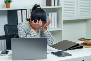 Asian office worker overwhelmed with work stress, holding head in hands at desk with laptop, papers, and files, showcasing realistic corporate challenges in a modern workspace.