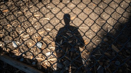 Aufkleber - A medium closeup of the workers shadow intermingles with tered debris as the sun filters through a chainlink fence creating an intricate pattern on the ground.