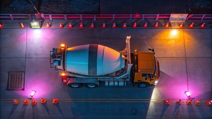 Poster - The drones camera focuses on a concrete mixer its rotating barrel catching the light while nearby fixtures and safety cones create a vibrant palette of colors in the nighttime