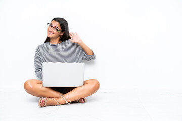 Wall Mural - Young caucasian woman with a laptop sitting on the floor saluting with hand with happy expression