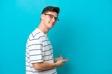 Wall Mural - Young handsome Brazilian man isolated on blue background extending hands to the side for inviting to come