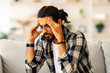 © Prostock-studio - Bearded indian guy in casual with glasses sitting on couch alone at home, touching his head and looking down, suffering from headache or experiencing difficulties with job, copy space