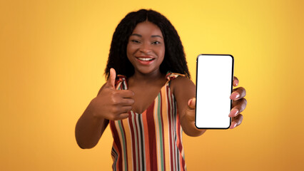 Lovely black woman demonstrating cellphone with empty black screen, showing thumb up, recommending new cool mobile shopping app on orange studio background, mockup for design