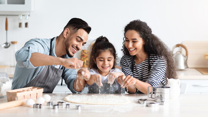 Wall Mural - Cheerful arab father, mother and daughter making biscuits together, rolling pastry dough and adding flour, middle-eastern mom and dad showing their little kid how to make cookies, kitchen interior