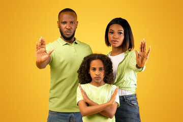 Protest, disagree and denial gesture. African american parents and daughter showing stop sign and girl crossed arms, standing on yellow background, studio shot