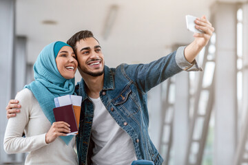 Wall Mural - Joyful Young Islamic Spouses Taking Selfie With Smartphone In Airport, Millennial Muslim Spouses Man And Woman In Hijab Holding Passports And Tickets, Ready For Honeymoon Trip, Travelling Together