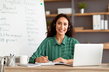 Aufkleber - Remote Study Concept. Portrait of smiling woman in wireless headset sitting at desk with pc laptop near whiteboard. Happy lady posing and looking at camera, writing, taking notes in paper notebook