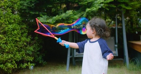 Wall Mural - A little Asian child enjoys Playing blowing Colorful soap bubbles Having Fun in a Lush Green Garden surrounded by greenery and sunlight on a Sunny Day, capturing the essence of carefree childhood fun.