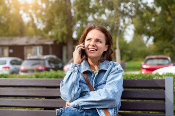 Wall Mural - Young woman wearing denim jacket sitting on a bench on a city street talking on a mobile phone.