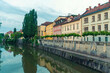 © Connect Images - A serene Ljubljana river flows past colorful buildings flanked by lush green trees under a soft blue sky at dusk, Ljubljana, Croatia