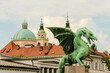 © Connect Images - A bronze dragon statue stands on Dragons Bridge in the foreground with the historic architecture of St. Nicholas Cathedral softly focused in the background under a cloudy sky, Ljubljana, Sloevnia