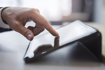 Poster - Close detail of a man's finger making contact with the screen of a sleek digital tablet resting on a desk, set against the soft-focus backdrop of a light-filled office