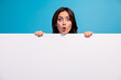 © deagreez - Surprised young woman peeking over white blank banner against blue background, with expressive astonishment and vibrant colors