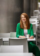 © Alexander - Young businesswoman with red hair in green pantsuit enjoying pause sitting at table in outdoor cafe. Urban atmosphere. Combination of professionalism, style and working time. Copy space.