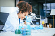 © NanSan - An African American female scientist in a white coat and blue gloves smiles confidently, holding a test tube with blue liquid. She conducts drug testing and medical research in a laboratory