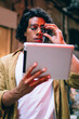© BullRun - Handsome pensive male in eyewear watching videos on digital tablet using application and 4G internet connection, trendy 20s man standing near neon light with touchpad blogging and share content