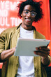 © BullRun - Cheerful handsome curly male in eyewear enjoying share and reading news on social networks via digital tablet, smiling 20s hipster guy standing outdoors near neon lights downloading application