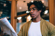 © BullRun - Serious concentrated male in eyewear spending time in cafe interior reading news from printed media on leisure, handsome curly man in casual wear holding newspaper checking financial information