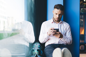 Fototapete - Caucasian male blogger sitting at windowsill and using 4g wireless connection on modern cellular technology, handsome millennial typing content text for sending sms message and post publication