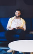 © BullRun - Portrait of cheerful Caucasian coworker resting at comfortable couch holding modern smartphone technology in hands and smiling at camera, happy male blogger with cellphone posing in loft interior