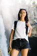 © BullRun - Cheerful female traveler with phone standing against waterfall
