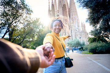 Wall Mural - POV of young mixed race couple holding hands in front of Sagrada Familia in Barcelona. Smiling girlfriend looking camera on street of city enjoying vacation happy and smiling. Funny tourism people