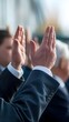 © Sarra - hands raised during a vote or business meeting. In the foreground, a raised hand is clearly visible wearing a navy blue suit sleeve with visible buttons,The image is taken in a professional setting