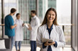 © fizkes - Beautiful female medical worker in white coat holding digital tablet device posing for camera with positive, joyful expression, standing in modern clinic skyscrapers office. Workflow, tech, medicine