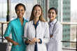 © fizkes - Group of three female doctors wear uniforms stand together in skyscraper clinic office, smiling at camera, exude amity, reliability, collaborative spirit of medical professionals. Medicine, teamwork