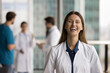 © fizkes - Laughing woman therapist wear white uniform looking at camera, standing in modern skyscraper clinic office, staff members blurred in background. Healthcare professional, head short portrait, success