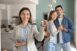© fizkes - Happy confident professional realtor woman showing key, looking at camera with toothy smile, posing for business portrait in apartment for rent or sale with couple of buyers, renters in background