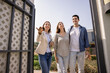 © fizkes - Positive real estate professional agent inviting couple of renters to come, standing at open fence door, pointing hand away. Realtor and customers standing at wicket gate, looking around