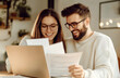 © Macrostock - Cheerful young couple using laptop and analyzing their finances with paper document. Smiling husband and wife using online banking service at home or receiving positive decision from bank by email