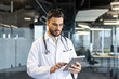 © Liubomir - Serious thinking male doctor working inside clinic office with tablet computer. Doctor using app for online consultation of patient remotely.