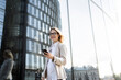 © muse studio - Professional woman in glasses smiles while using smartphone near modern building in city