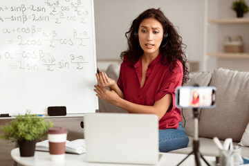 Aufkleber - E-Learning. Confident woman having video conference call using laptop. Lady teacher wearing wireless headphones talking to webcam, recording tutorial with mobile phone on tripod, streaming live