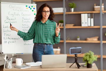 Aufkleber - Distant Education. Portrait of young lady teacher in glasses and earbuds standing at whiteboard, explaining English grammar rules to students. Lady recording lesson using cellphone on tripod