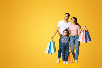 Wall Mural - Shopping Offer. Smiling Arab Family With Little Daughter Carrying Shopper Bags And Pointing Aside At Copy Space Over Yellow Background, Happy Parents And Female Kid Enjoying Seasonal Sales, Panorama