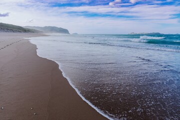 Wall Mural - Tranquil beach scene with gentle waves lapping at the shore. Footprints in the sand lead to the ocean. Peaceful coastal view. Opoutere, Whangamata, Coromandel Peninsula, New Zealand