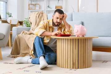 Wall Mural - Stressed young man with calculator and piggy bank counting money at home