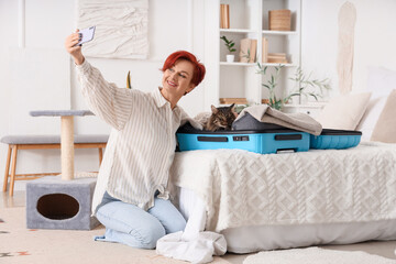 Wall Mural - Happy mature woman taking selfie with cute cat in open suitcase at home