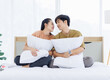 © Bangkok Click Studio - Portrait of young lovely sweet attractive asian happy couple love posing to camera smiling hugging pillow sitting on white bed in bedroom at home good time romantic together Valentine's day concept.