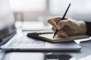 Poster - Sharp view of a woman's hand transcribing in a notepad on a cutting-edge laptop, with a defocused backdrop