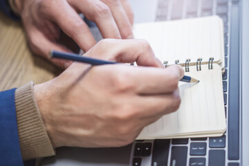 Poster - A man's hand is caught in close-up, busily writing in a notepad on a laptop, while the background is out of focus