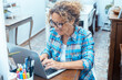 © simona - Confident woman at home wearing glasses and curly hair, smiling while working remotely on her laptop, managing online projects, conducting web research, or engaging in social media activities