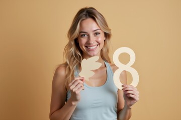 Smiling woman holding a leaf and the number eight against a soft yellow background during a creative photoshoot in a modern studio